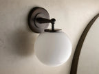 Close-up of the antique bronze Fulton Sconce with a glowing milk glass globe attached to a round backplate.
