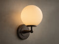 Front view of the Antique Bronze Fulton Sconce, featuring a lit milk glass spherical globe, mounted on a textured gray wall.