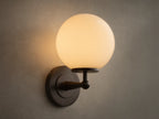Front view of the Antique Bronze Fulton Sconce, featuring a lit milk glass spherical globe, mounted on a textured gray wall.