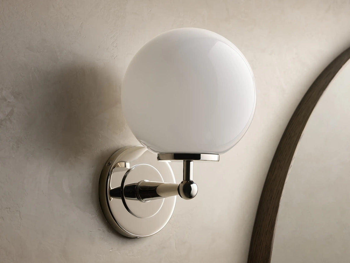 Front view of the polished nickel Fulton Sconce with a glowing spherical milk glass globe, mounted on a wall.