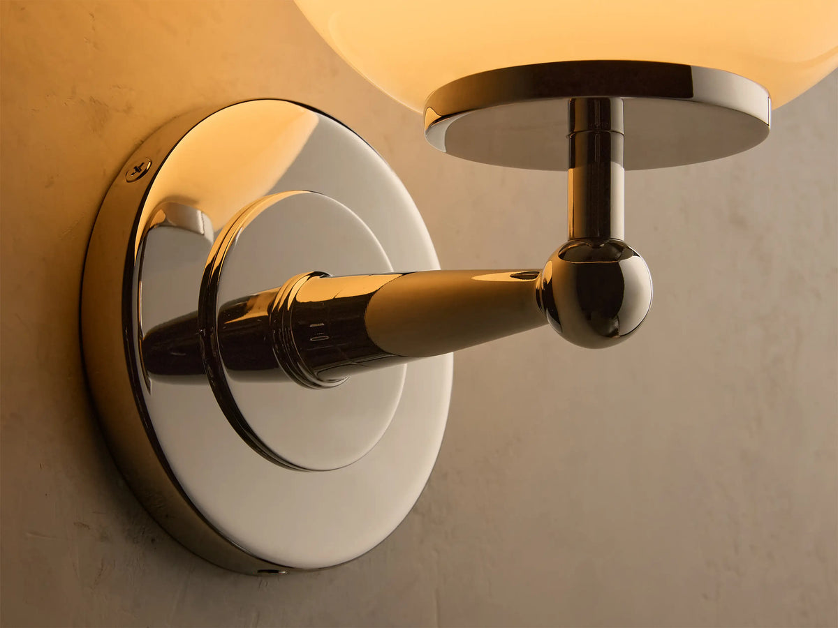 Front view of the polished nickel Fulton Sconce with a round milk glass globe, mounted on a textured light gray wall.