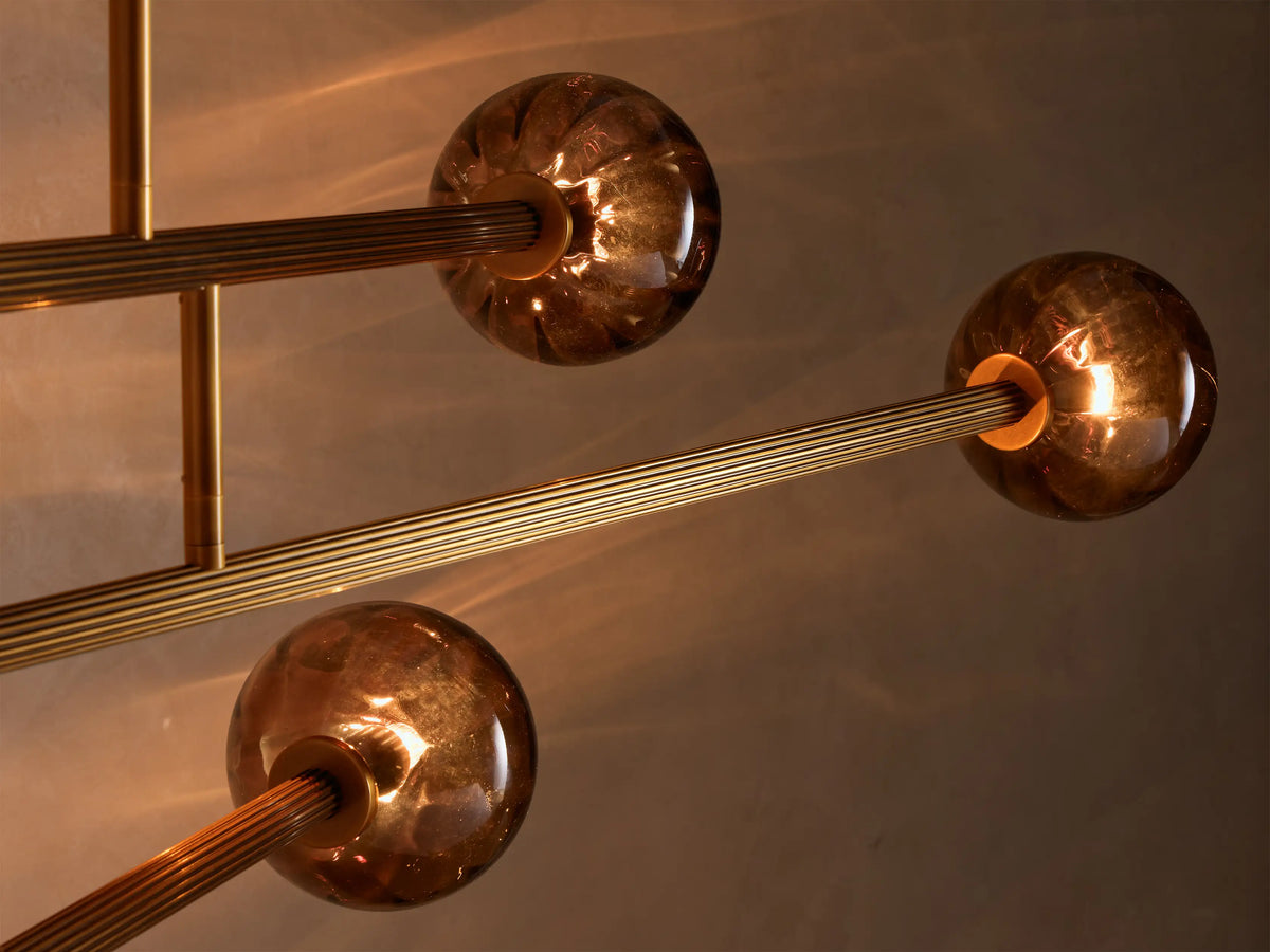 Close-up of a modern light fixture featuring three glowing amber spherical glass shades and ribbed brass-colored rods.