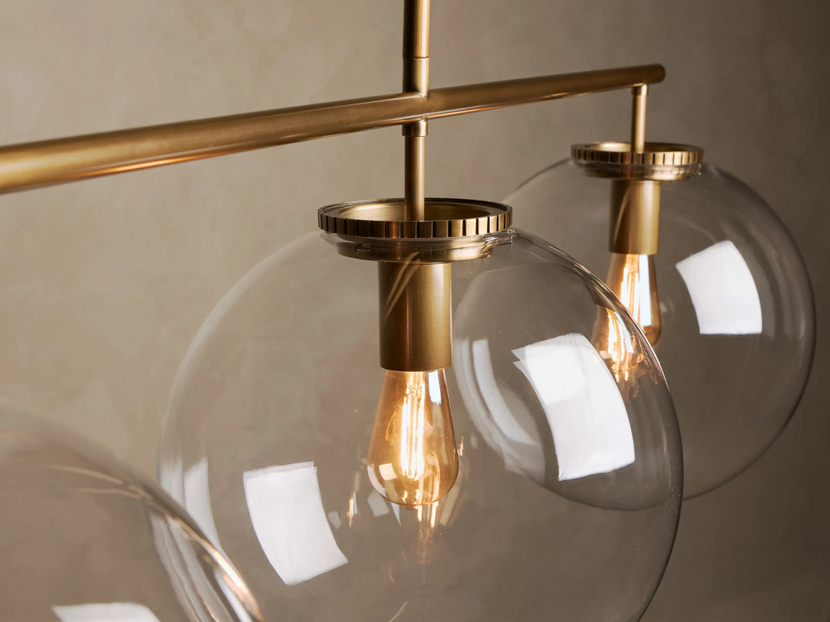 Front view of a linear pendant light featuring three clear glass globe shades, brass hardware, and exposed filament bulbs.