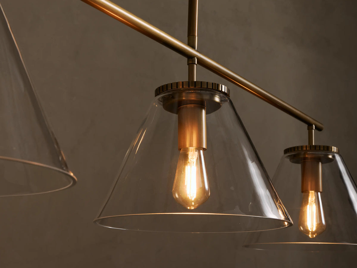 Front view of a brass-finished Three-Light Linear Chandelier with three clear conical glass shades and exposed Edison-style bulbs.
