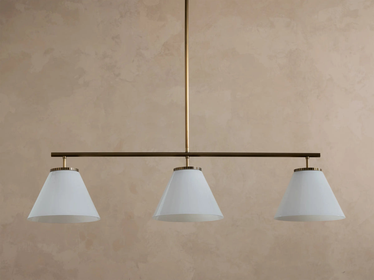 Front view of three-light linear chandelier with white conical shades, a dark bronze bar, and a brass suspension rod, emitting a warm glow.
