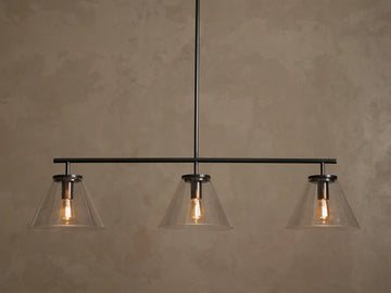 Front view of a black linear pendant light fixture with three clear conical glass shades and exposed warm Edison bulbs.