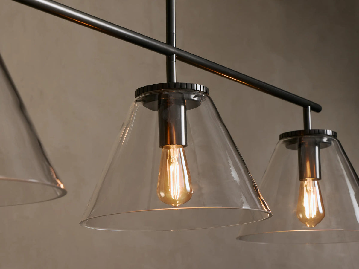 Front view of a black metal linear pendant light with three clear glass cone shades and amber Edison bulbs.