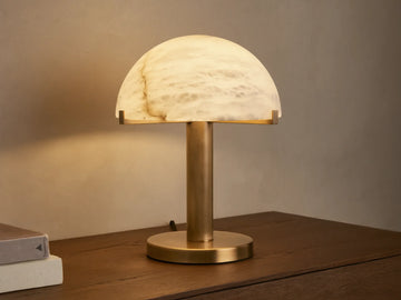 Emerie Table Lamp with alabaster stone shade and brass base on a wooden table, softly lit. Black fabric cord visible.