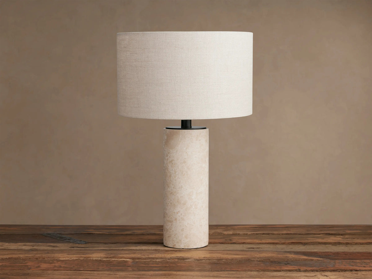 Brixton Table Lamp: Cylindrical light marble base, dark stem, and beige linen-texture drum shade on a rustic wood surface.
