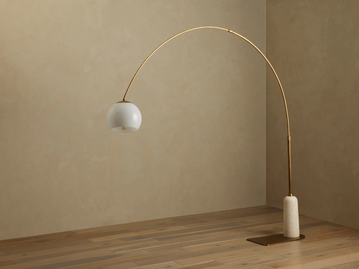 Christie Floor Lamp: arched brass-finished steel, spherical milk glass shade, and natural stone base on wood floor.