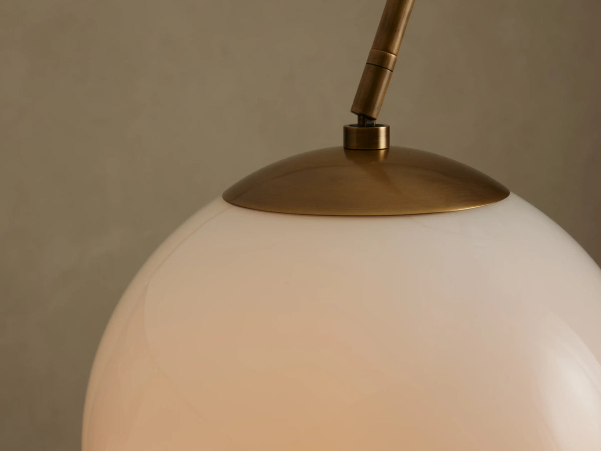 Close-up of Christie Floor Lamp's glowing milk glass shade with a brass cap and arm extending up-left, against a neutral wall.