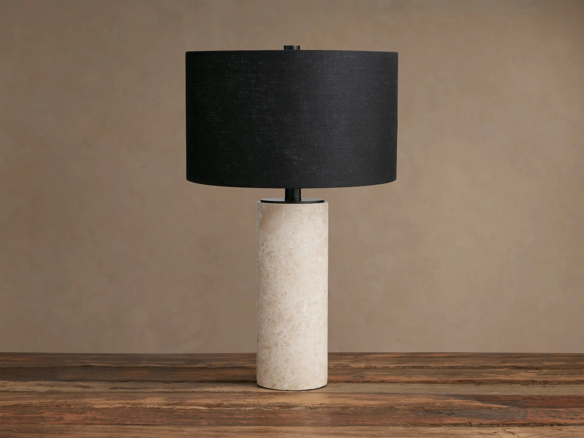 Brixton Table Lamp with cylindrical natural marble base and black fabric drum shade, standing on a rustic wooden table.