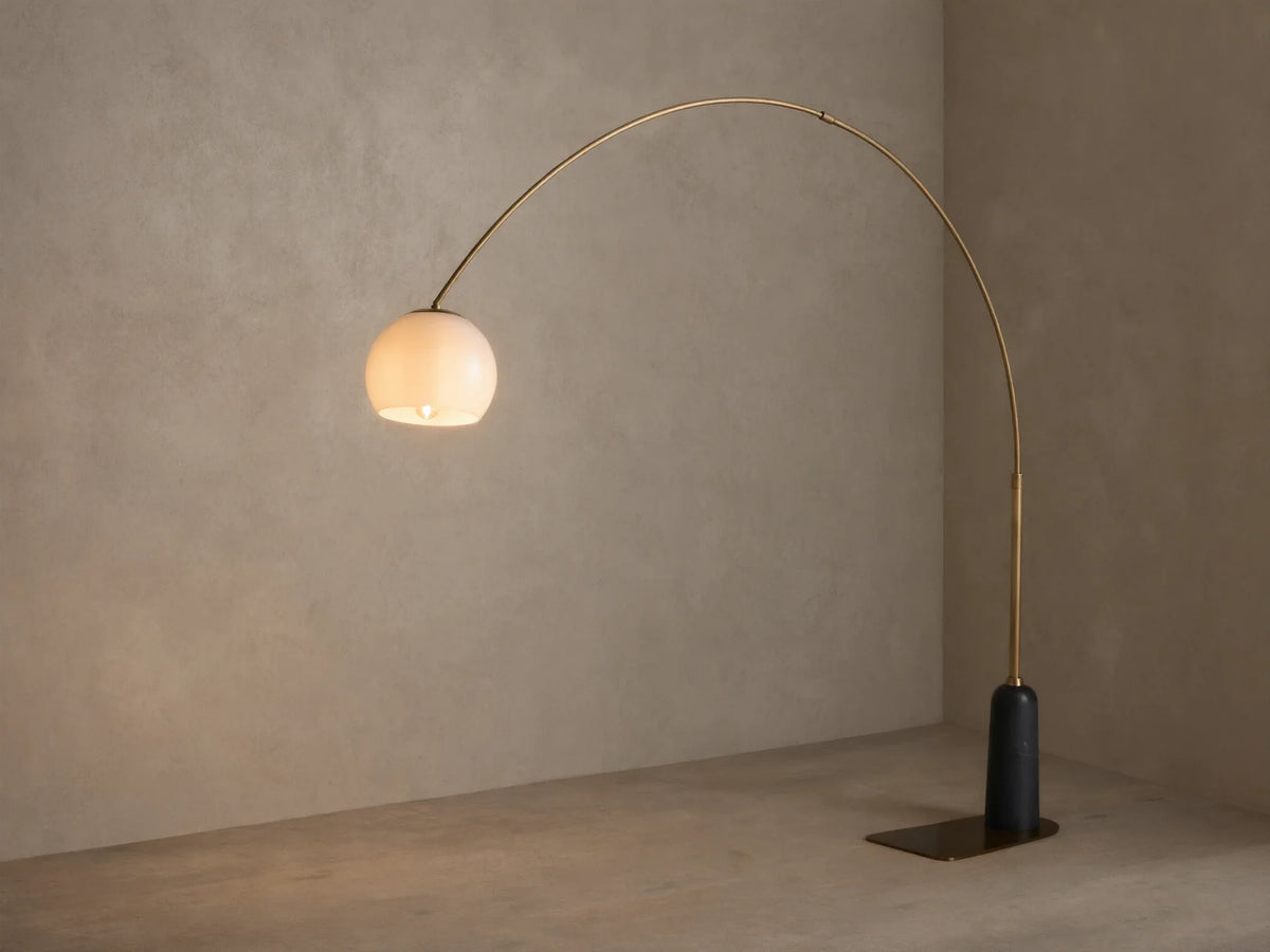 Christie Floor Lamp: Arched brass-finished lamp with glowing milk glass shade, dark stone base on wood floor against beige wall.