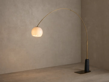 Christie Floor Lamp: Arched brass-finished lamp with glowing milk glass shade, dark stone base on wood floor against beige wall.