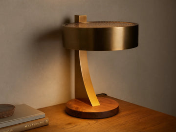 Alastair Table Lamp with a burnished brass cylindrical shade, perforated top, curved arm, and oiled walnut base, emitting warm light on a wooden table.
