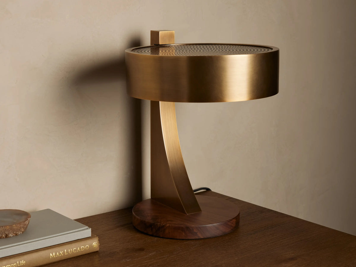 Alastair Table Lamp with a burnished brass cylindrical shade, perforated top, curved arm, and oiled walnut base, emitting warm light on a wooden table.