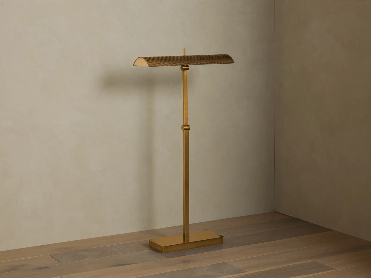 Abel Metal Floor Lamp: Upright, brass finish, rectangular base, long perforated shade emitting warm LED light. Beige wall, wood floor.