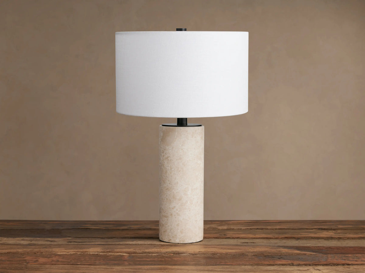 Brixton Table Lamp, front view. Cylindrical light beige marble base, dark metal stem, white fabric drum shade, on wood.