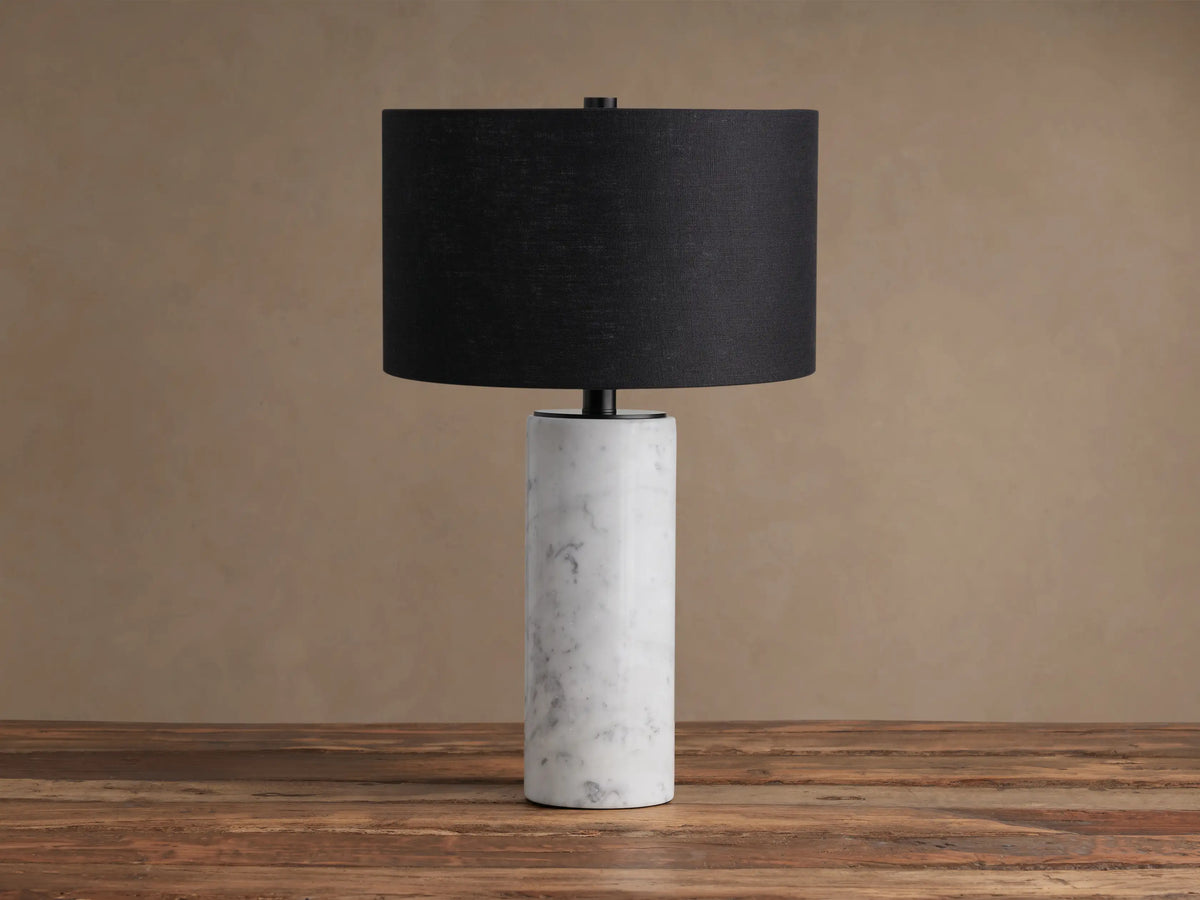 Brixton Table Lamp with a white cylindrical marble base and a black fabric drum shade, resting on a rustic wooden table.