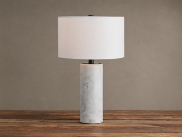 A Brixton Table Lamp featuring a cylindrical white marble base, black stem, and white fabric shade on a wooden table.