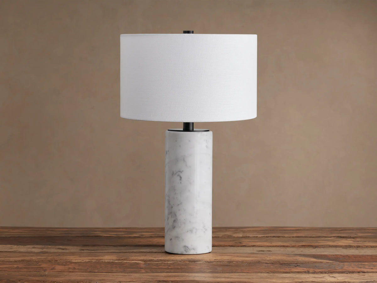 A Brixton Table Lamp featuring a cylindrical white marble base, black stem, and white fabric shade on a wooden table.