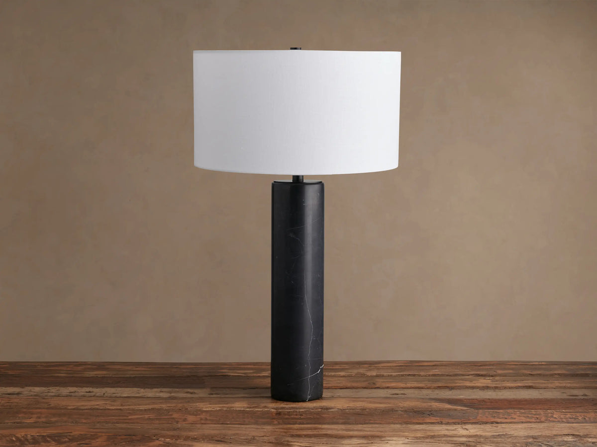 Brixton Table Lamp with a tall, cylindrical black marble base & white drum shade, standing on a rustic wooden table.