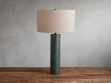 Brixton Table Lamp with a cylindrical green marble base and a large cream fabric lampshade, placed on a wooden surface against a gray wall.