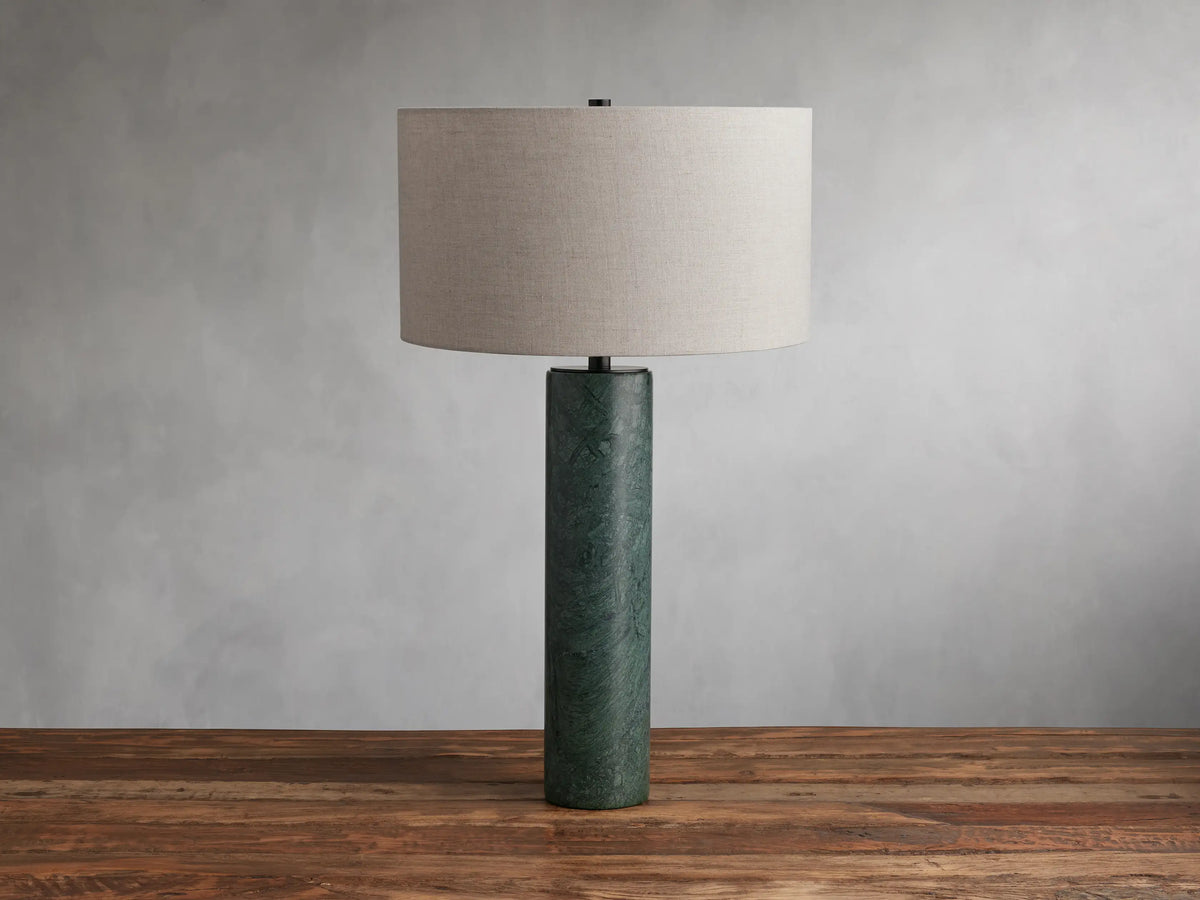Brixton Table Lamp with a cylindrical green marble base and a large cream fabric lampshade, placed on a wooden surface against a gray wall.