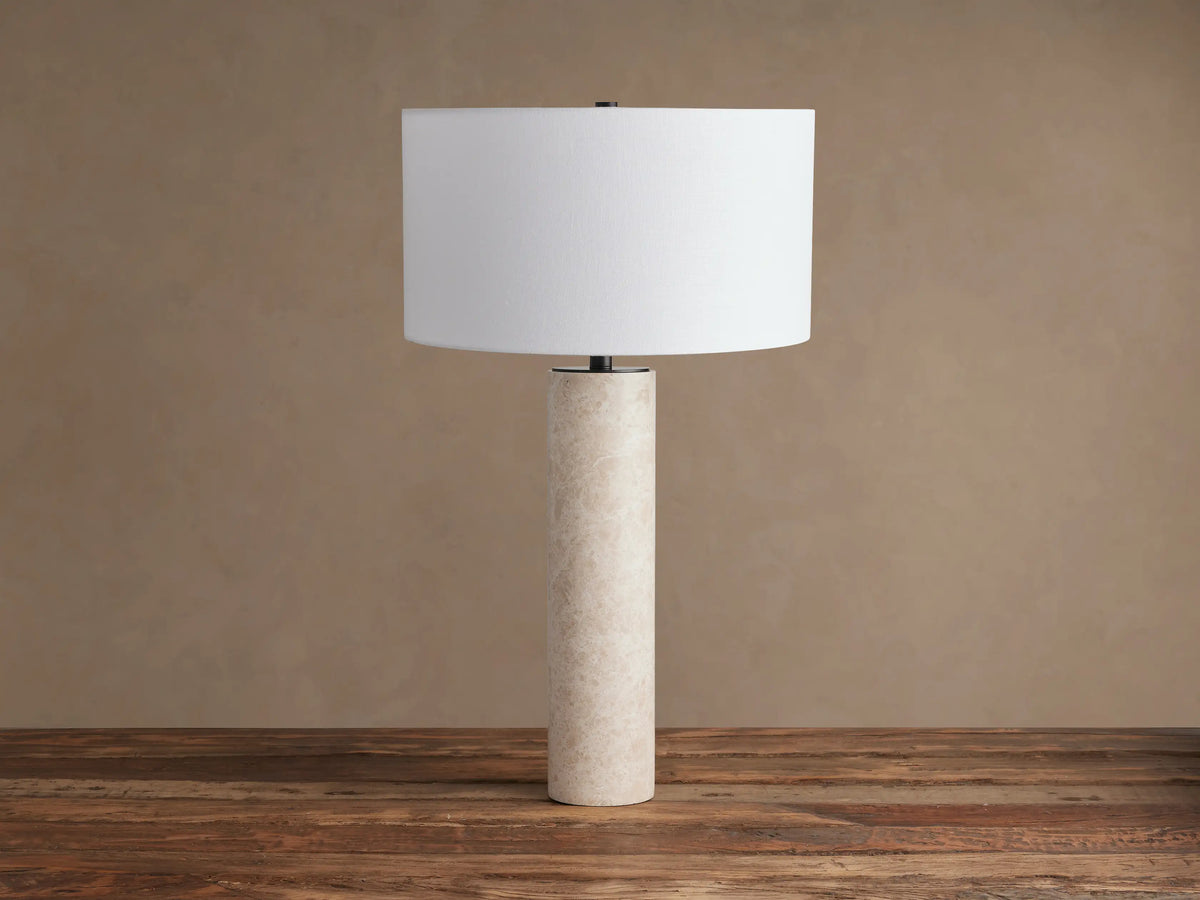 Brixton Table Lamp with a tall, cylindrical light beige marble base and a crisp white drum shade on a rustic wood table.
