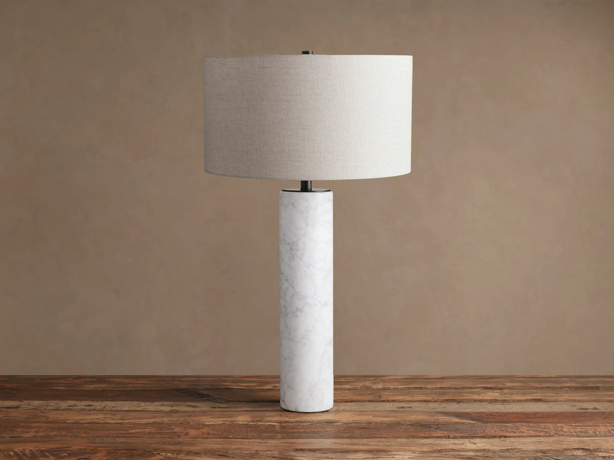 Brixton Table Lamp, a cylindrical white marble base, dark metal neck, and light grey fabric shade on a rustic wood surface.