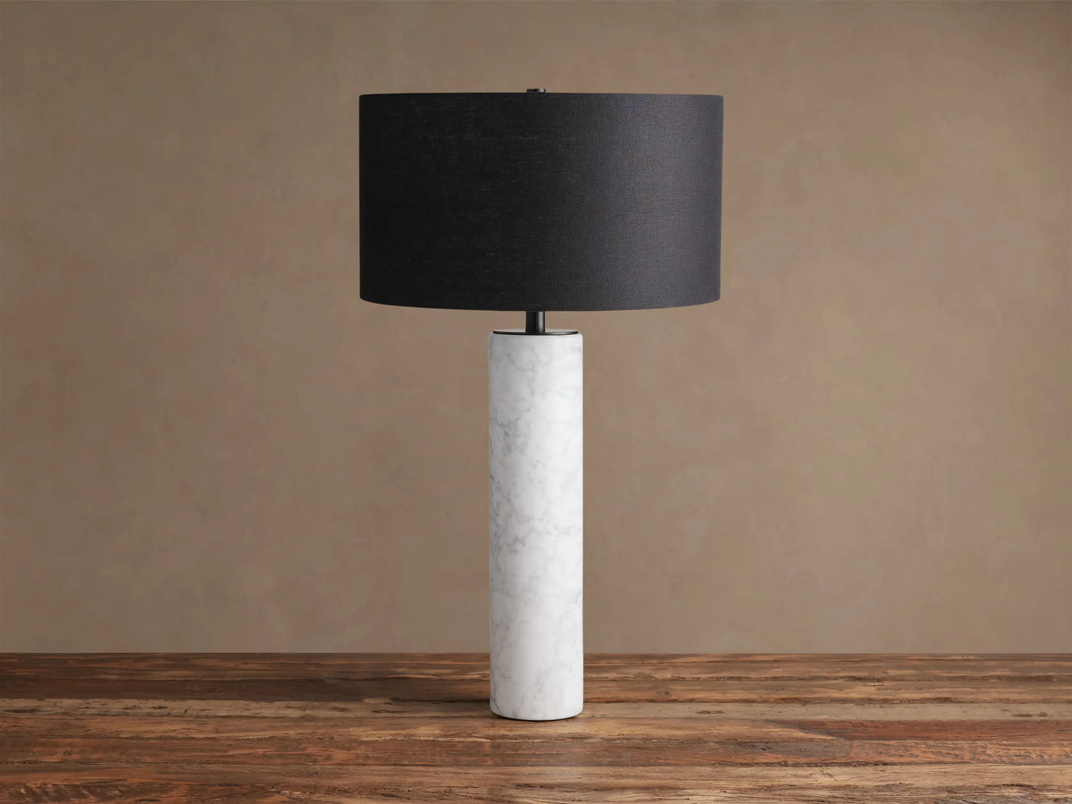 Brixton Table Lamp featuring a tall, cylindrical white marble base with gray veins and a black fabric drum shade, on a wood surface.