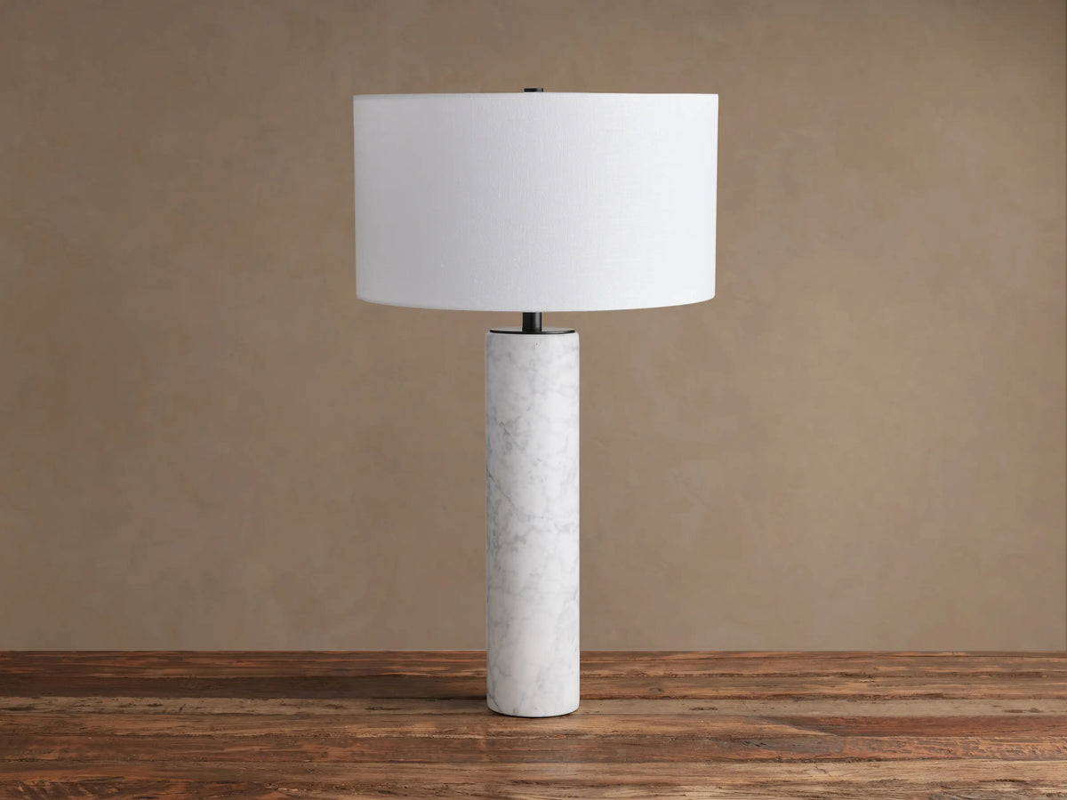Brixton Table Lamp with a white marble cylindrical base, dark stem, and white drum shade on a rustic wooden table.