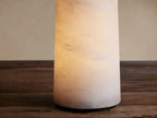 Kelby Outdoor Rechargeable Lamp, a white alabaster cylinder with subtle marbling, on a distressed wooden table against a tan wall.