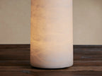White cylindrical Kelby Outdoor Rechargeable Lamp made of alabaster with natural patterns, standing on a dark wooden surface.