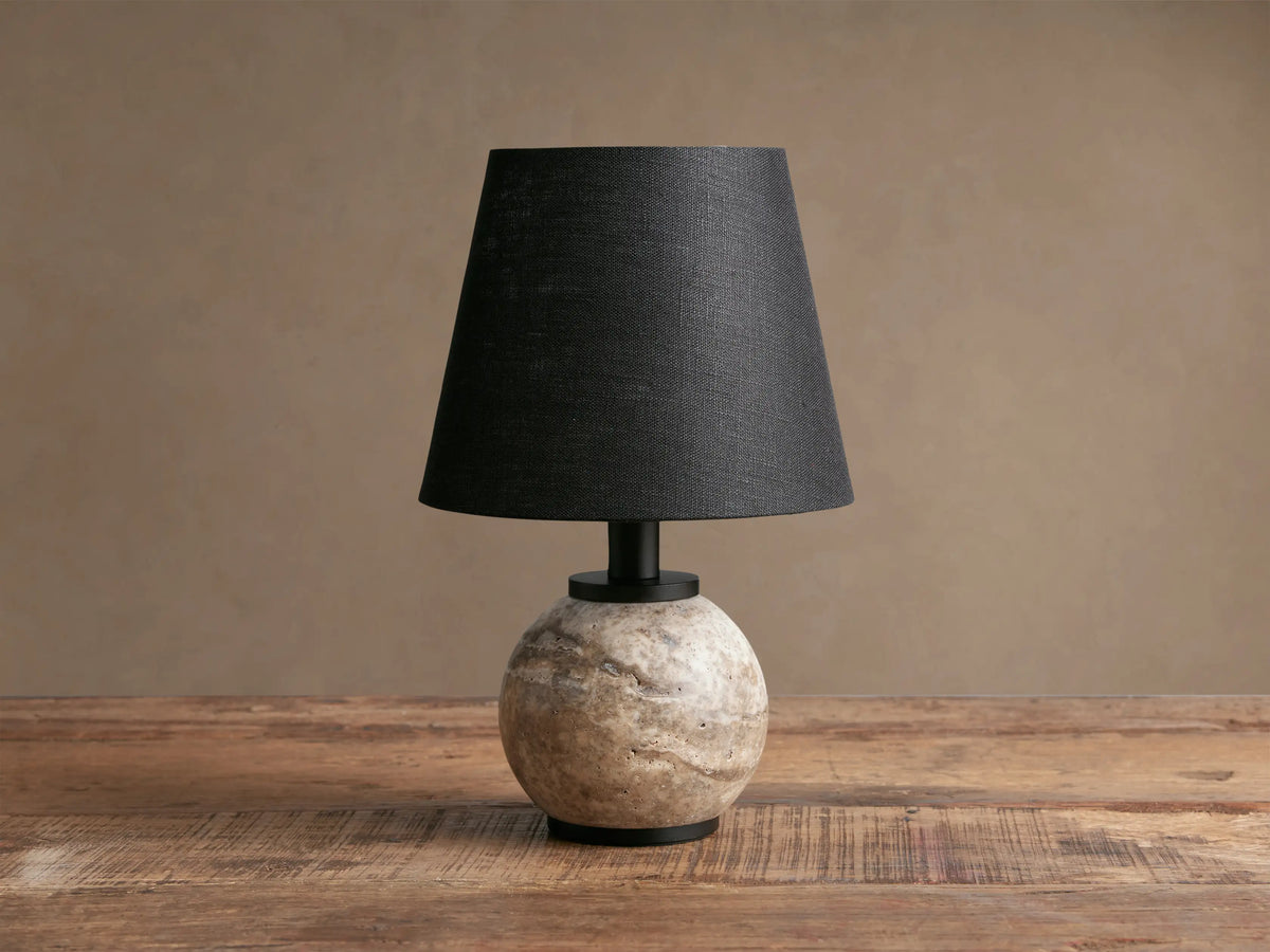 Celia Outdoor Rechargeable Lamp with a spherical cream/grey travertine base, black linen shade, and matte black hardware on a rustic wooden table.