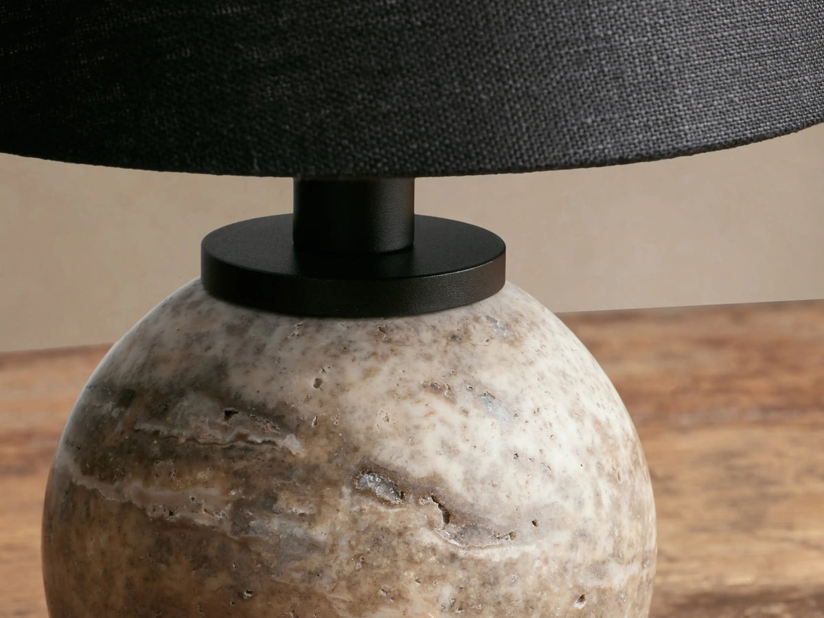 Celia Outdoor Rechargeable Lamp with a rounded, textured beige travertine base and a black conical linen shade on a wood table.