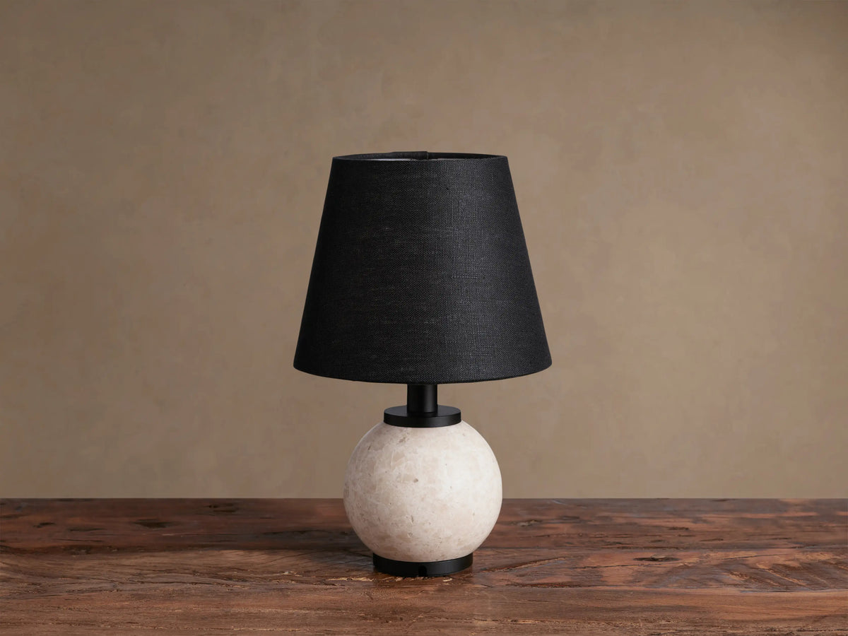 Celia Outdoor Rechargeable Lamp with a round cream marble base, matte black hardware, and a black linen shade on a wooden table.