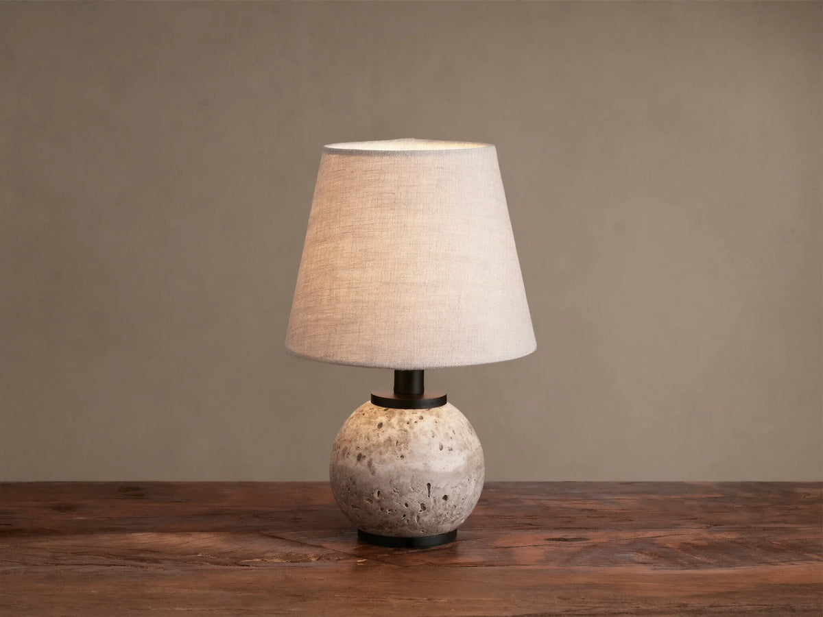 Celia Outdoor Rechargeable Lamp with a round, textured travertine base, matte black hardware, and an illuminated flax linen shade.