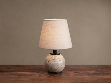 Celia Outdoor Rechargeable Lamp with a round, textured travertine base, matte black hardware, and an illuminated flax linen shade.