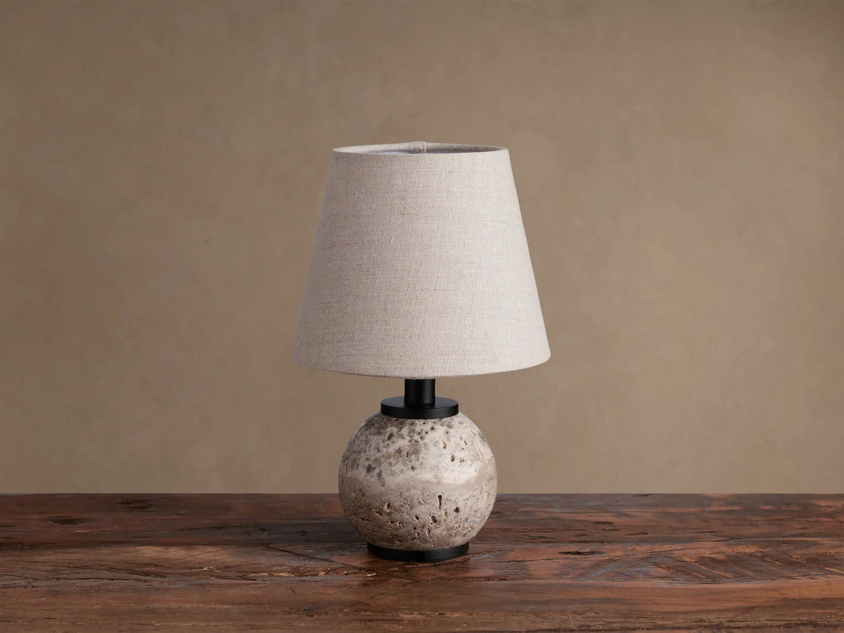 Celia Outdoor Rechargeable Lamp with a round, textured travertine base, matte black hardware, and an illuminated flax linen shade.