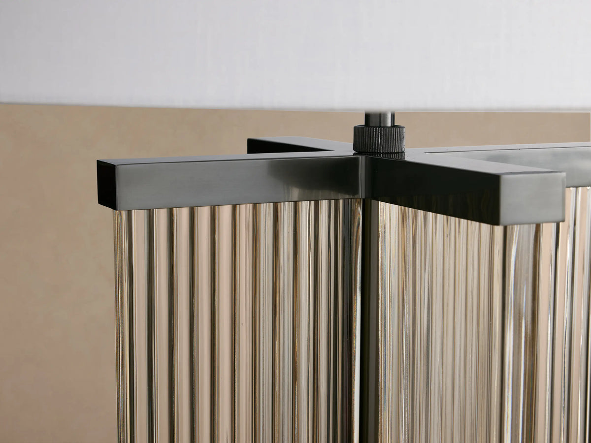 Delsie Table Lamp with a white cylindrical shade and a dark bronze steel base encasing fluted, smoky glass, on a wood table.