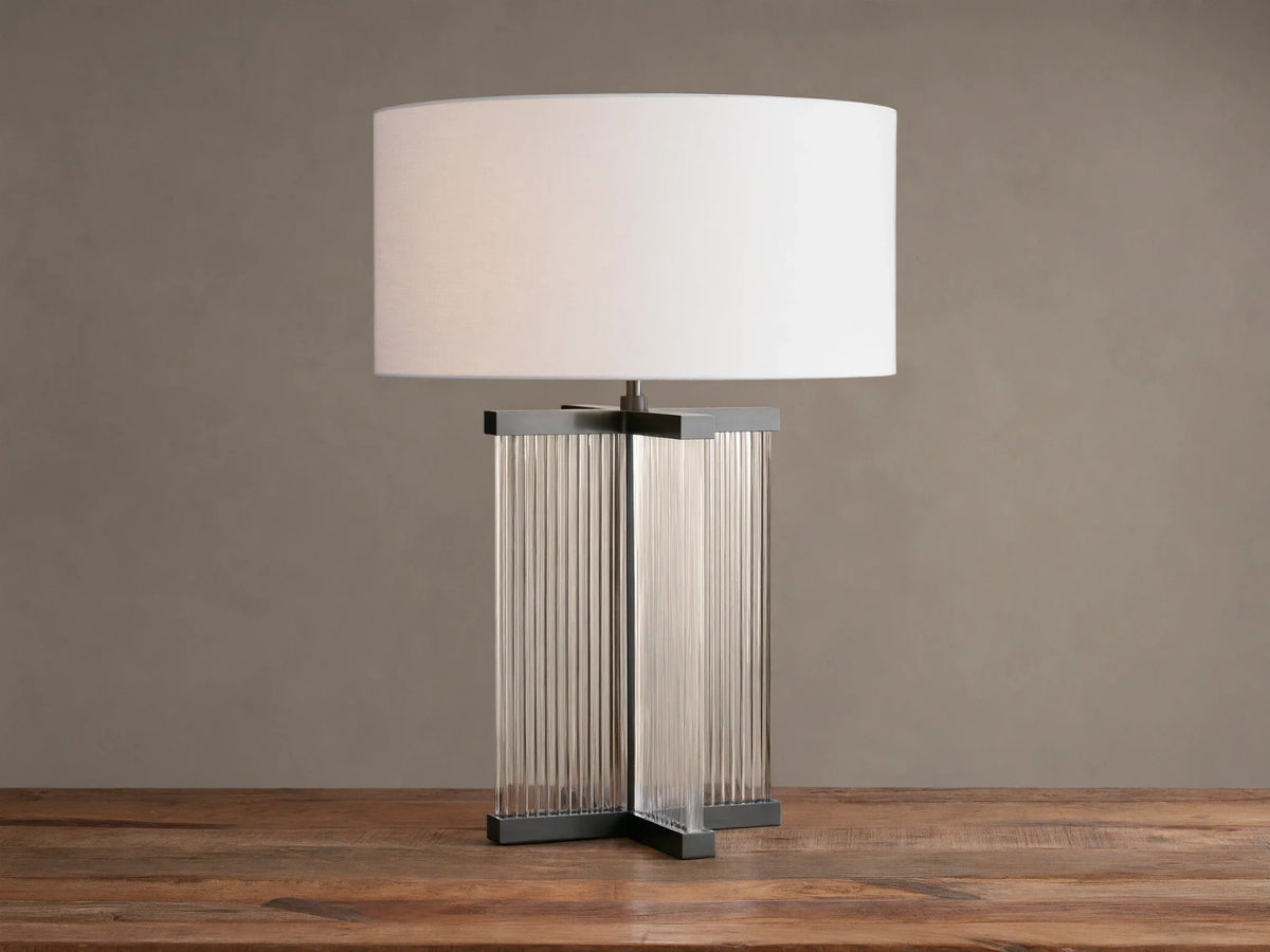 Delsie Table Lamp with a white drum shade over a dark metal base with clear fluted glass panels, on a rustic wooden table.