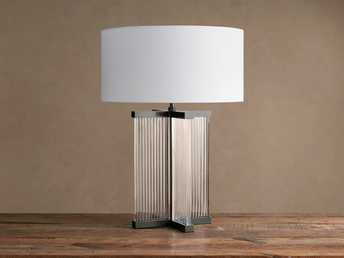 Delsie Table Lamp with a white drum shade over a dark metal base with clear fluted glass panels, on a rustic wooden table.