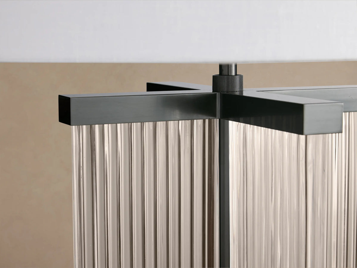 Delsie Table Lamp with a clear ribbed glass base, dark metal accents, and a white drum shade, on a wooden table.