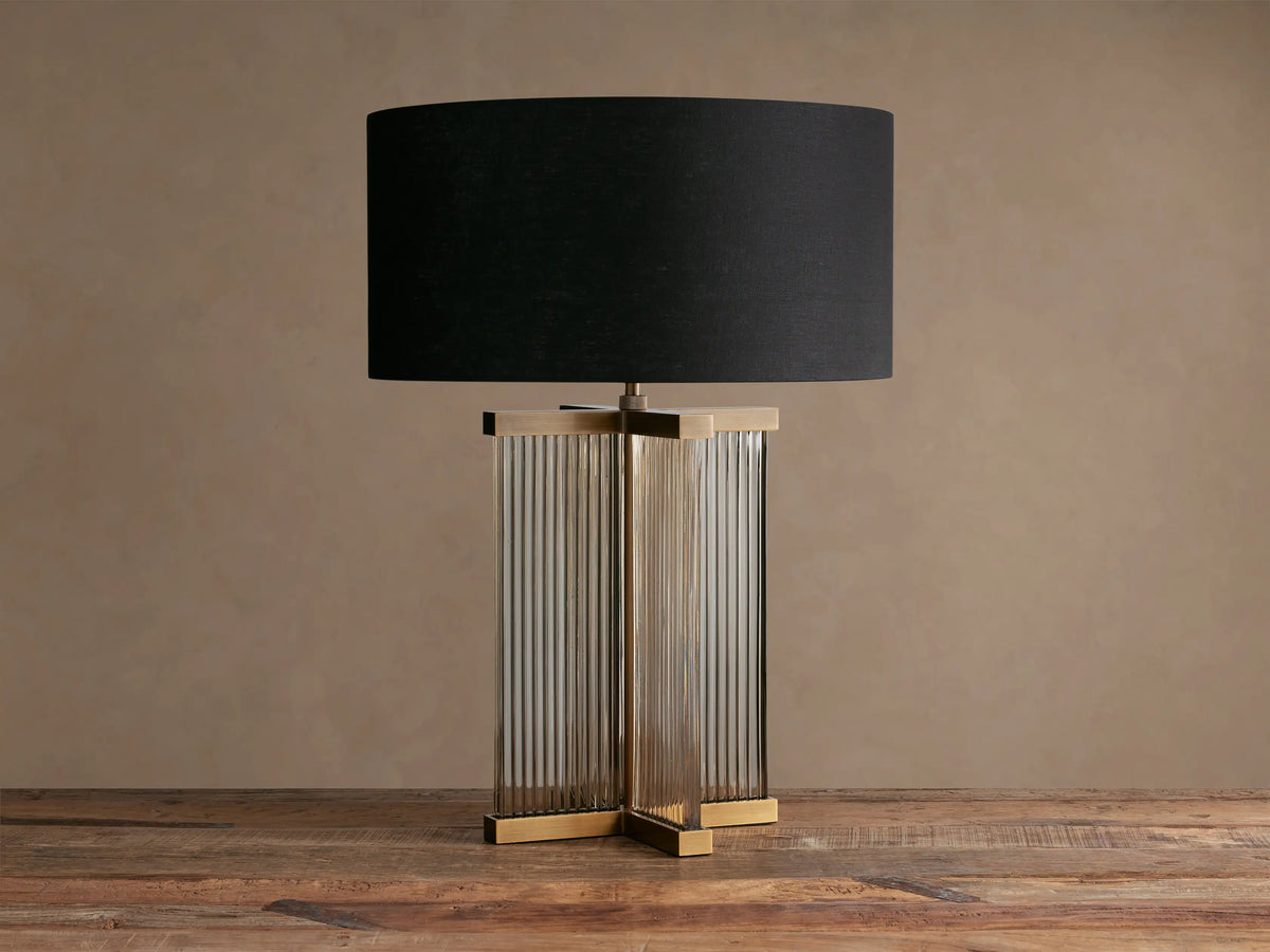 Delsie Table Lamp with black shade, fluted clear glass column base, and burnished brass top and bottom accents on a wooden table.