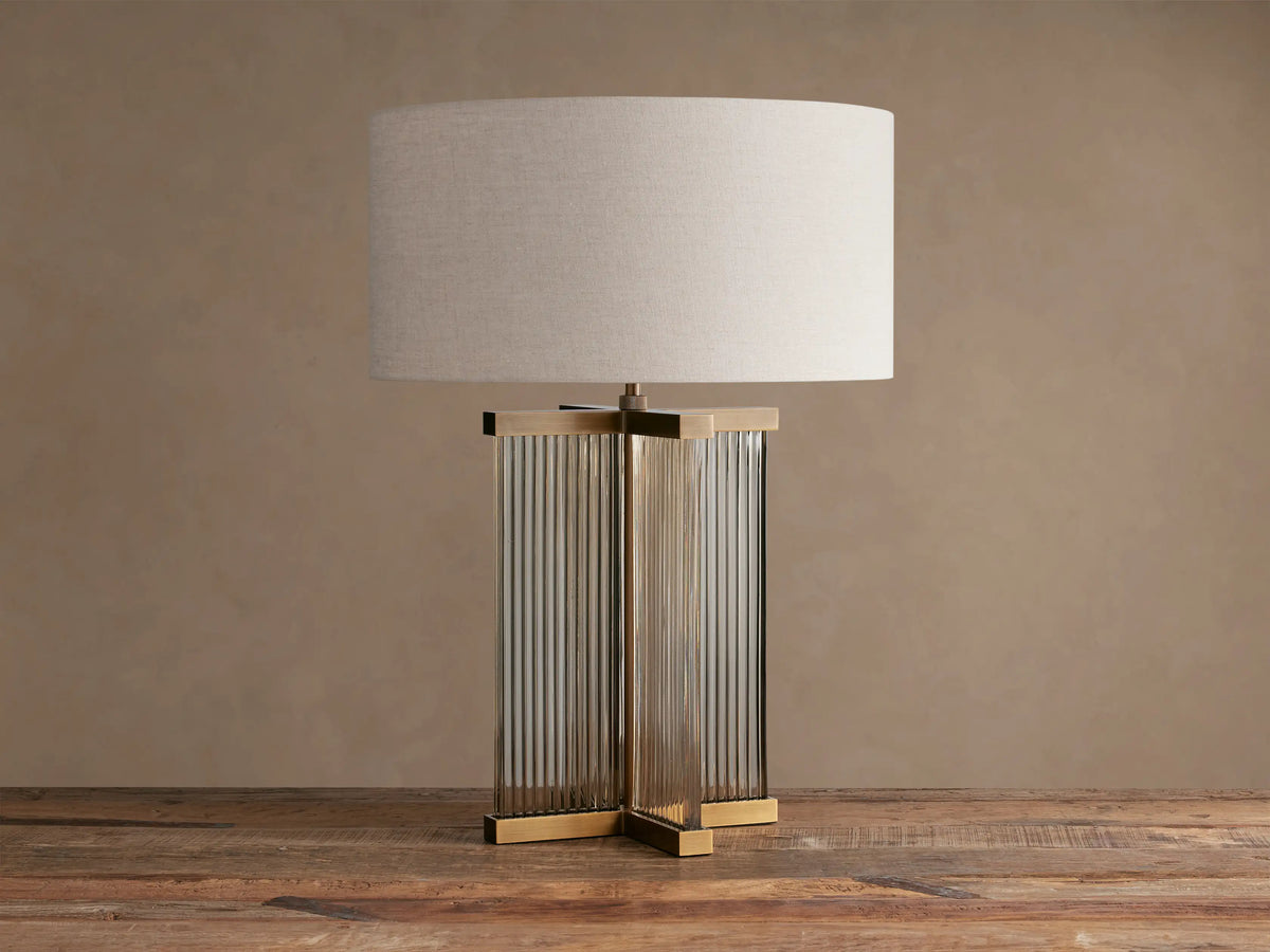 Delsie Table Lamp with a light flax drum shade, burnished brass frame, and ribbed glass base on a rustic wooden table.