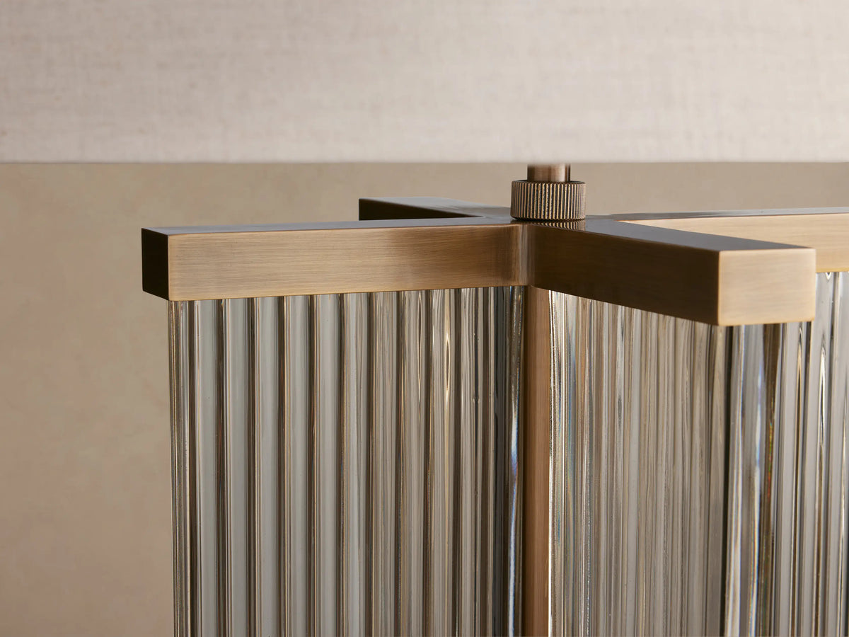 Delsie Table Lamp with a light flax cylindrical shade and a fluted clear glass base with burnished brass accents, on a wooden table.