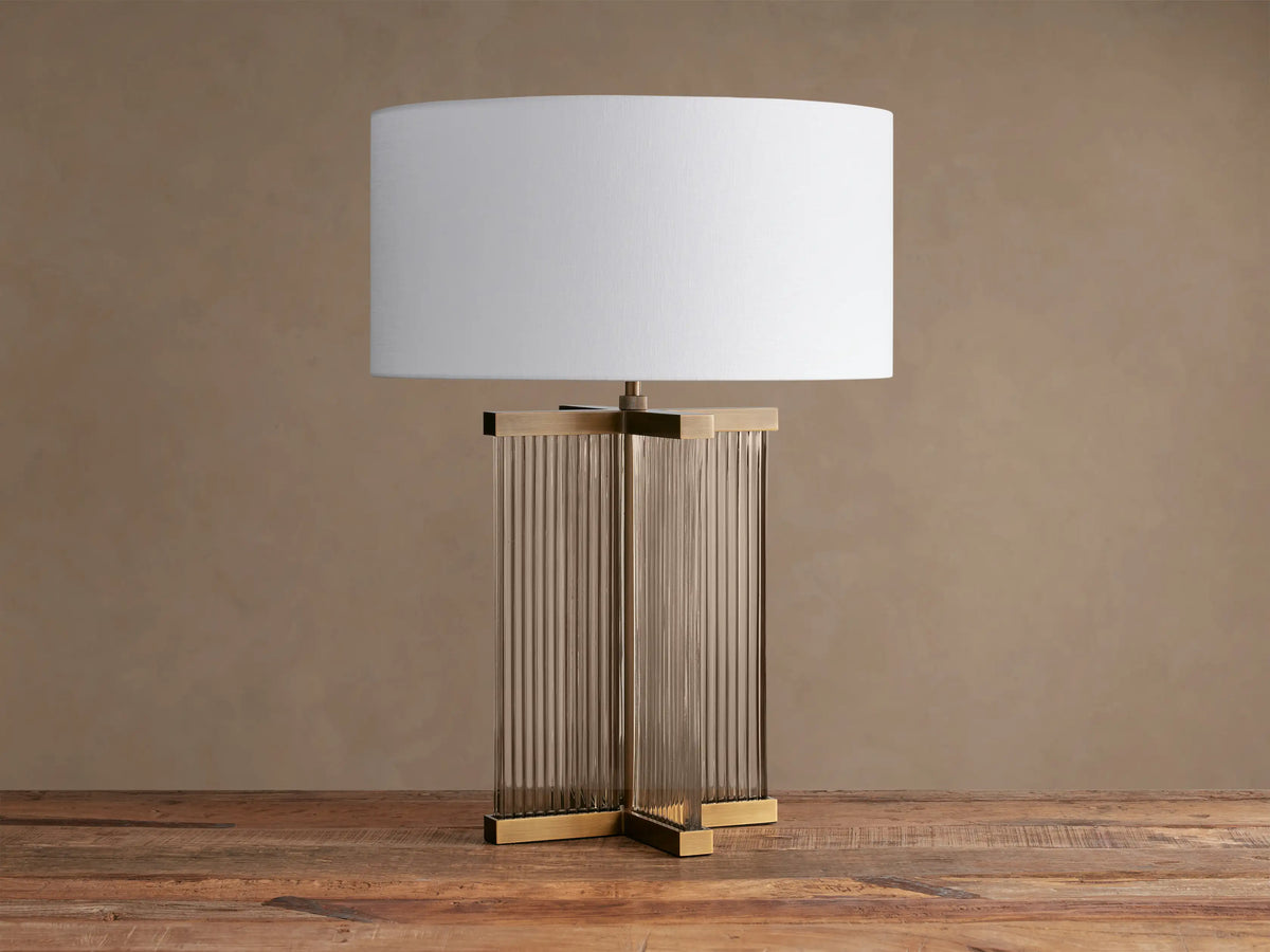 Delsie Table Lamp, white fabric shade atop a fluted glass and burnished brass base, sitting on a rustic wooden surface.