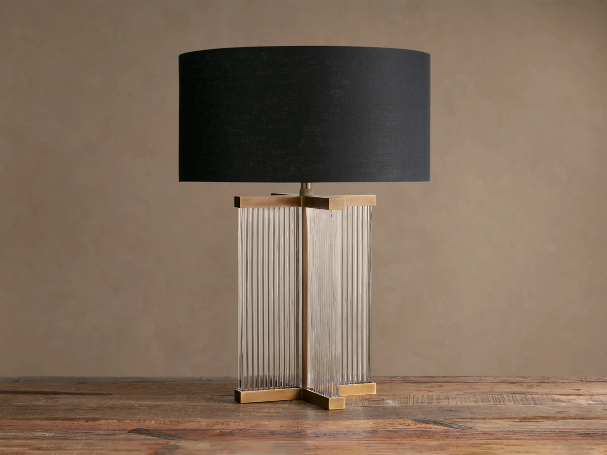 Delsie Table Lamp with a black drum shade, clear fluted glass rectangular base, and burnished brass accents on a wooden table.