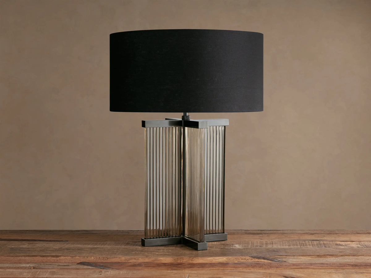 Delsie Table Lamp with a black fabric drum shade, fluted clear glass body, and burnished bronze metal base on a wood table.